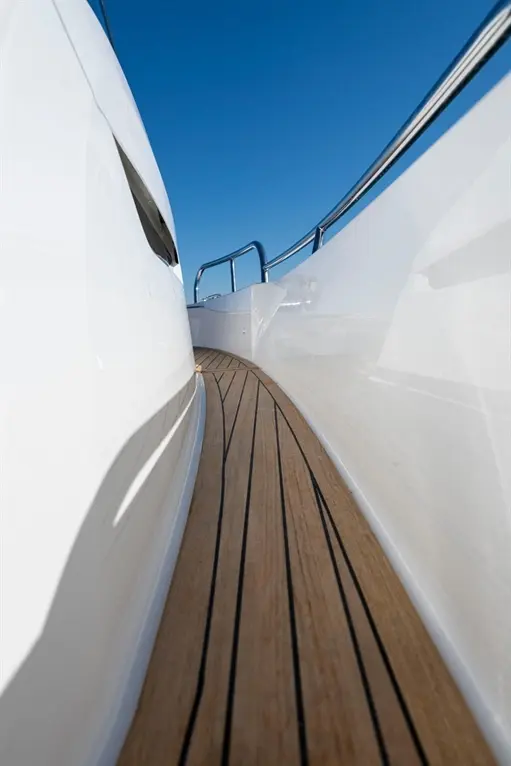 Close-up view of a yacht's polished wood deck with black stripes, surrounded by white hull and blue sky above.